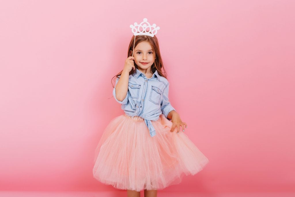 Cheerful little girl with long brunette hair in tulle skirt holding princess crown on head isolated on pink background. Celebrating brightful carnival for kids, birthday party, having fun cute kid.