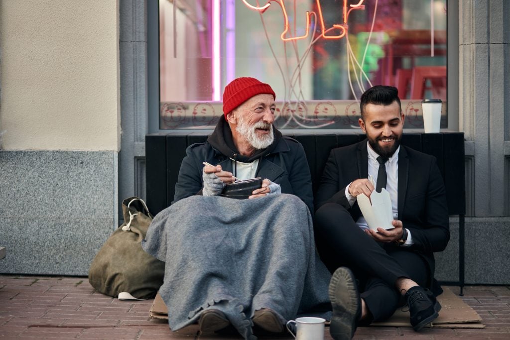 Smiling rich and poor men together sitting on street and eat while speaking. Happy despite social inequality