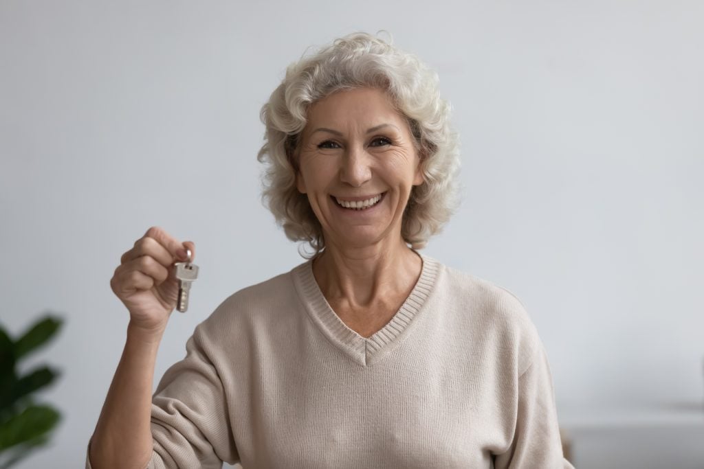 Happy old woman holds bunch of keys standing indoors smile look at camera. New house buying relocation, accommodation for holidays rent. Affordable low-income housing for seniors older adults concept