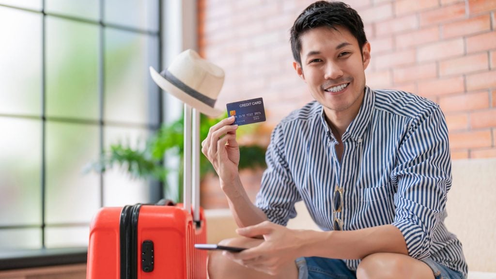 Ready to travel abroad,asia adult male man casual cloth sit on sofa couch next to luggage travel bag, hand hold credit card smile cheerful confident carefree face expression in hotel lobby new journey
