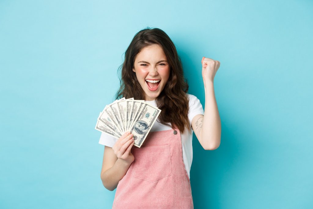 lucky young woman looks excited holding dollars celebrating