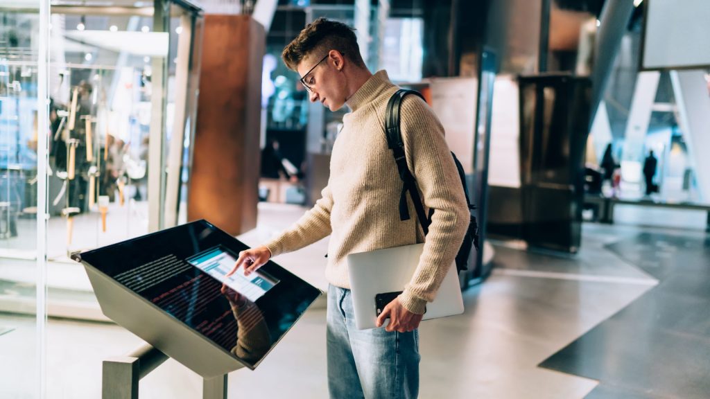 Side view of handsome young man in glasses and with backpack standing with gadgets in hands while using information panel in shopping centre