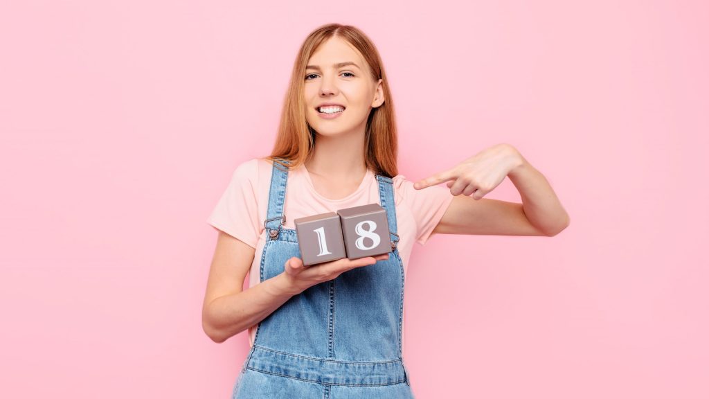 A cheerful cute young woman is celebrating her birthday and holding cubes with her age 18 eighteen years old, on an isolated pink background
