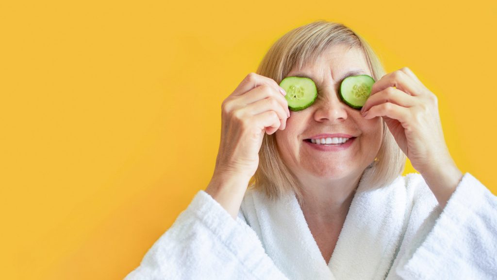 Cheerful senior woman with cucumber slices covering her eyes in front of her eyes on a yellow background. Spa beauty treatments at home, organic cosmetics, body care concept. Natural spa treatment.