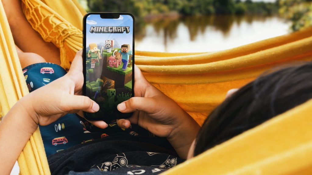 Child lying in hammock with Minecraft mobile game app on smartphone screen. Lake in the background. Rio de Janeiro, RJ, Brazil. May 2022.