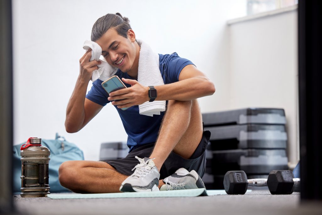 Fitness, phone or man with sweat on social media at gym in training, workout or exercise resting on break. Mobile app, digital or tired athlete relaxing on cellphone searching for body goals posts
