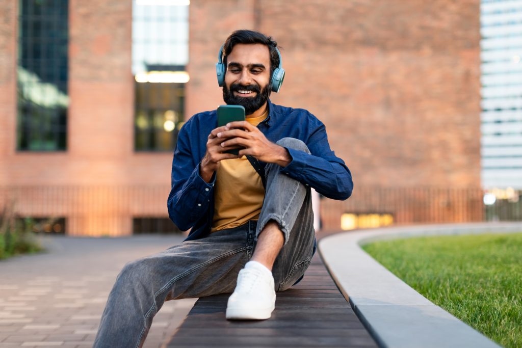 Indian man with headphones using phone enjoying music sitting outside