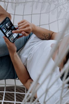 Faceless woman using a smartphone to browse music apps while relaxing on a hammock indoors.